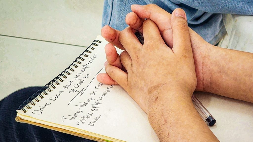 A close-up shows a person's clasped hands resting on a notebook. The page highlights concerns about online child protection, alongside a pen. The tone is contemplative.