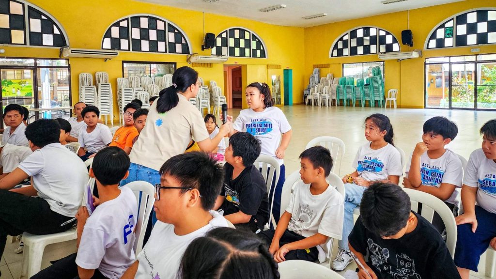 A diverse group of children and a woman engage in conversation in a bright room with yellow walls. Stacks of chairs and large windows are in the background.