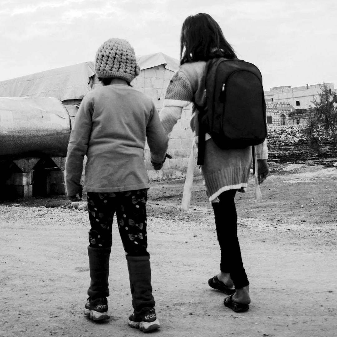 Two children, one wearing a backpack and the other a knit hat, hold hands while walking on a dirt road in a rural, desolate setting. The mood is somber.