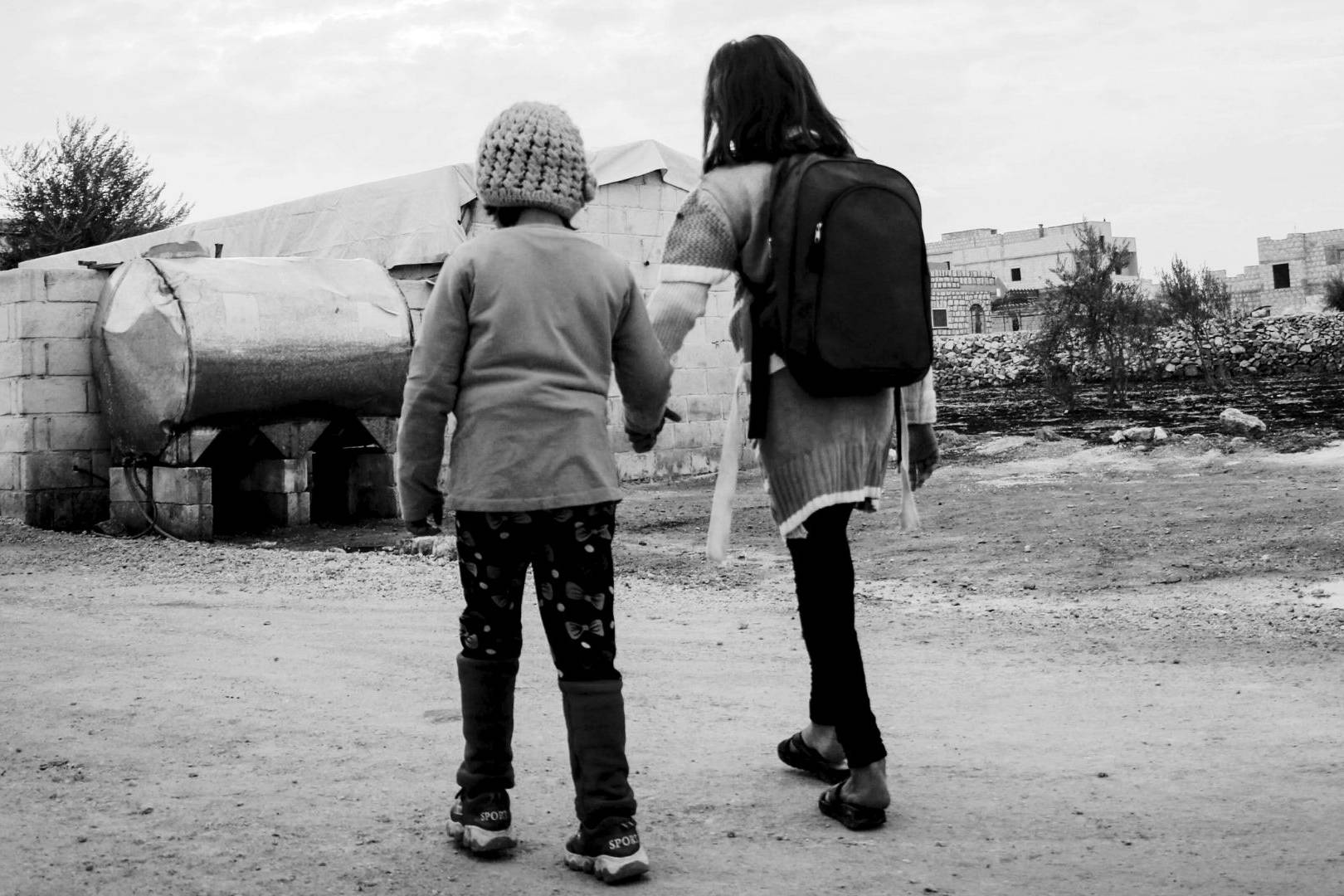 Two children, one wearing a backpack and the other a knit hat, hold hands while walking on a dirt road in a rural, desolate setting. The mood is somber.