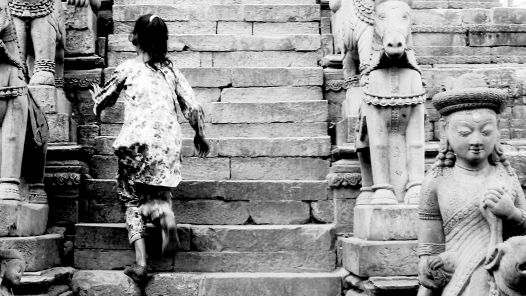 A young child runs up stone steps flanked by intricate statues of deities and animals. The black-and-white photo conveys a sense of exploration and history.