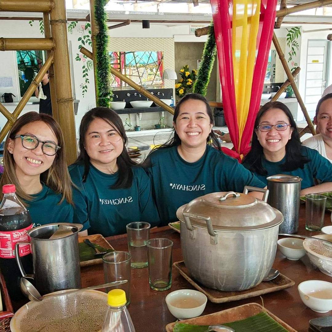 A group of eight people smiling around a dining table with empty plates, soda bottles, and a large pot. The mood is cheerful and relaxed.