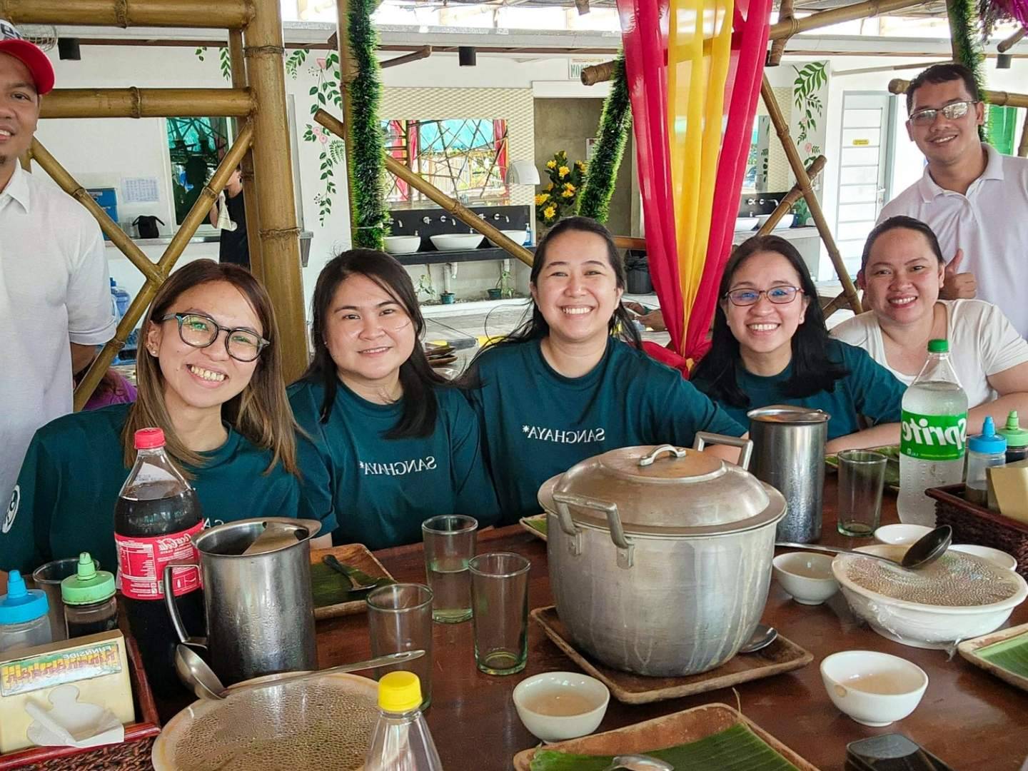 A group of eight people smiling around a dining table with empty plates, soda bottles, and a large pot. The mood is cheerful and relaxed.