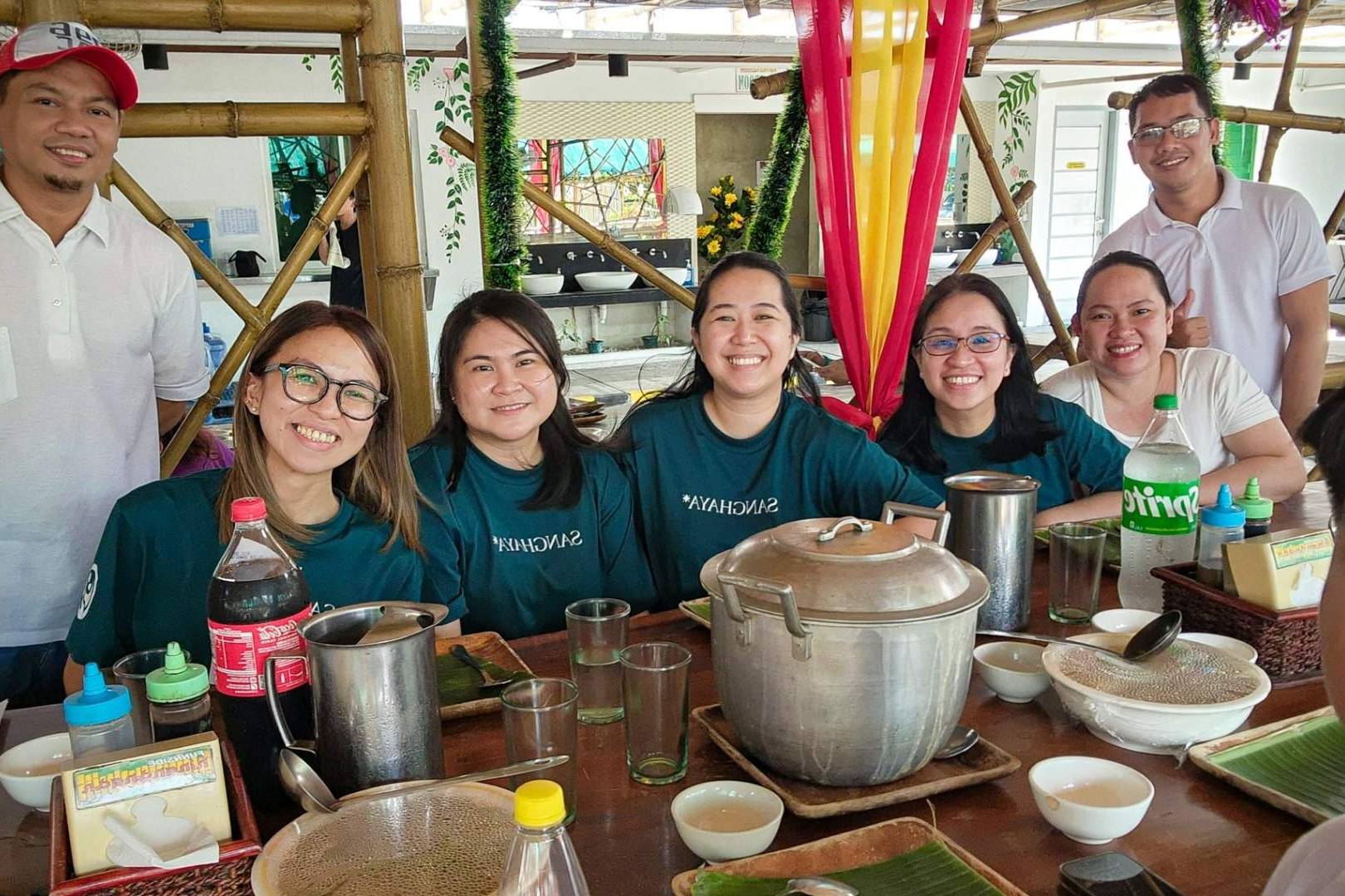 A group of eight people smiling around a dining table with empty plates, soda bottles, and a large pot. The mood is cheerful and relaxed.