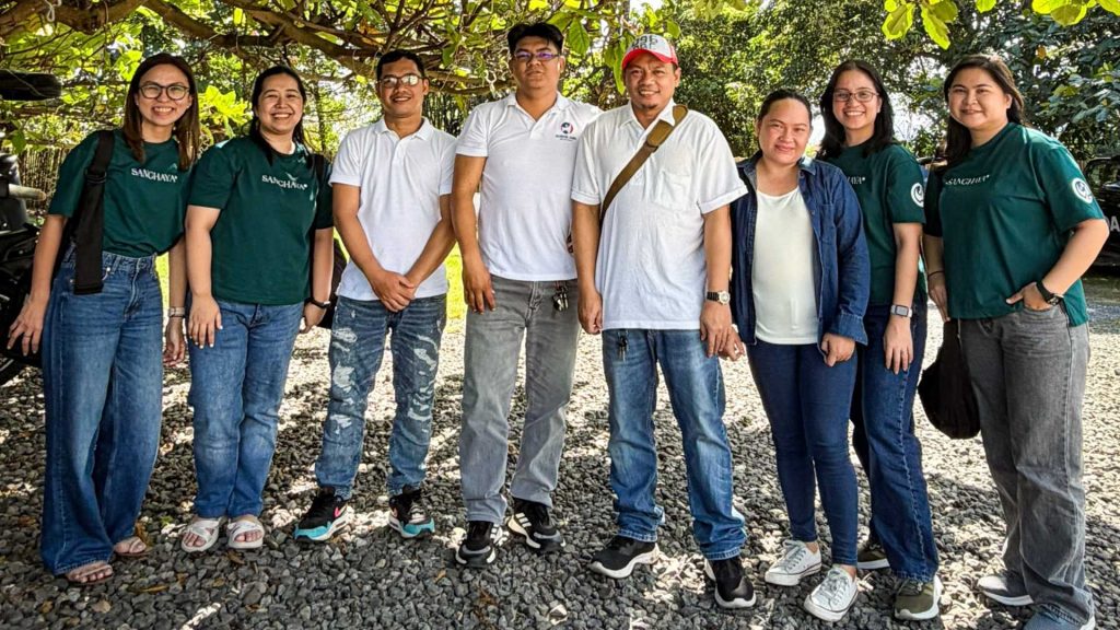 A group of eight people stand smiling outdoors under trees, wearing casual attire with green and white shirts, conveying a cheerful and relaxed atmosphere.