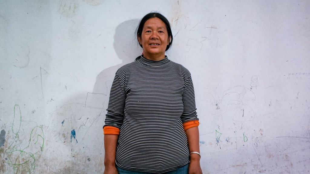 Woman in a striped sweater stands against a textured wall with faint drawings. She smiles warmly, creating a friendly and casual atmosphere.