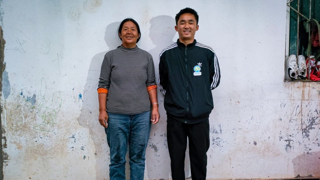 Two people stand smiling against a worn, white wall. The person on the left wears a striped shirt, while the person on the right is in a black jacket. Nearby, a window ledge holds several pairs of shoes. The mood is casual and friendly.