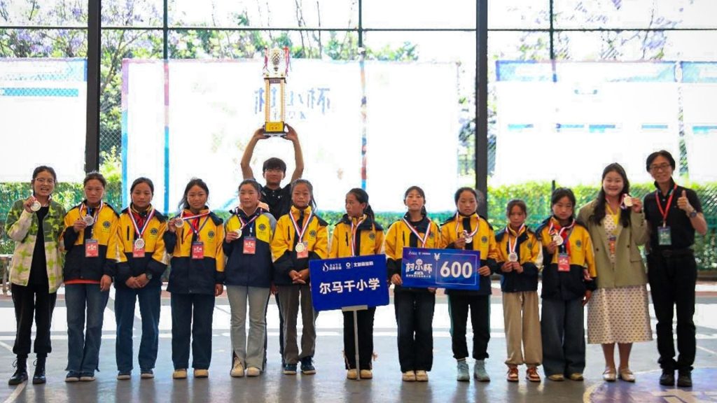 A group of young students in yellow jackets stand together indoors, celebrating a victory. They hold medals and trophies, smiling proudly.