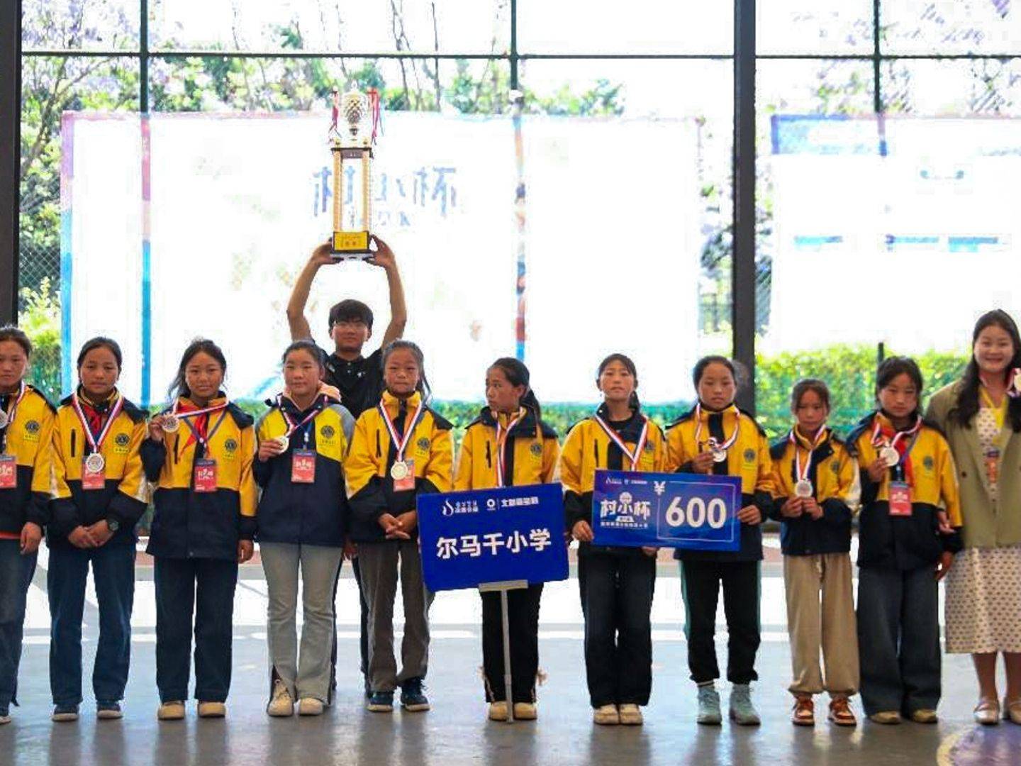 A group of young students in yellow jackets stand together indoors, celebrating a victory. They hold medals and trophies, smiling proudly.