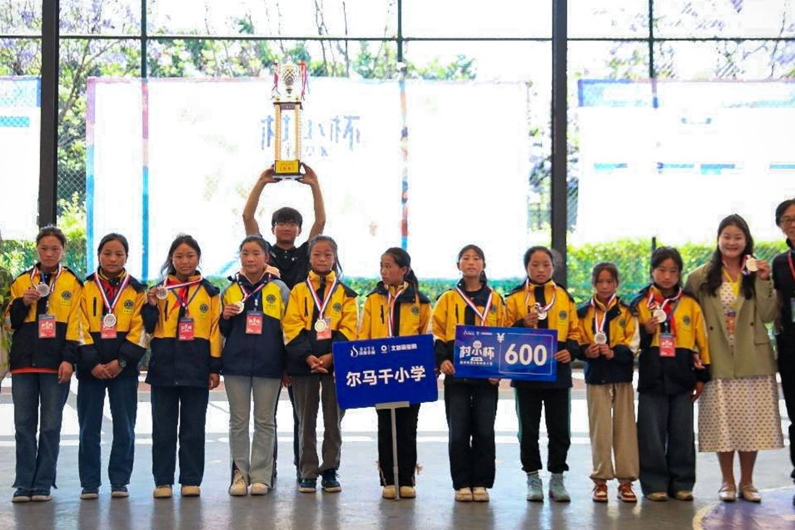 A group of young students in yellow jackets stand together indoors, celebrating a victory. They hold medals and trophies, smiling proudly.