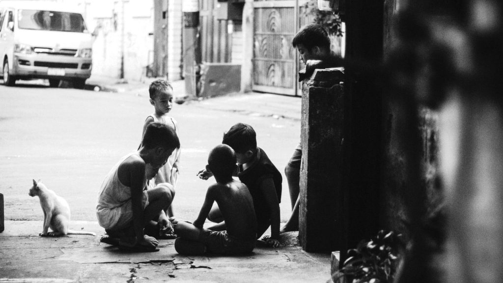 Children and a cat gather on a dimly lit urban street, engrossed in play. The black-and-white setting conveys a sense of simplicity and camaraderie.