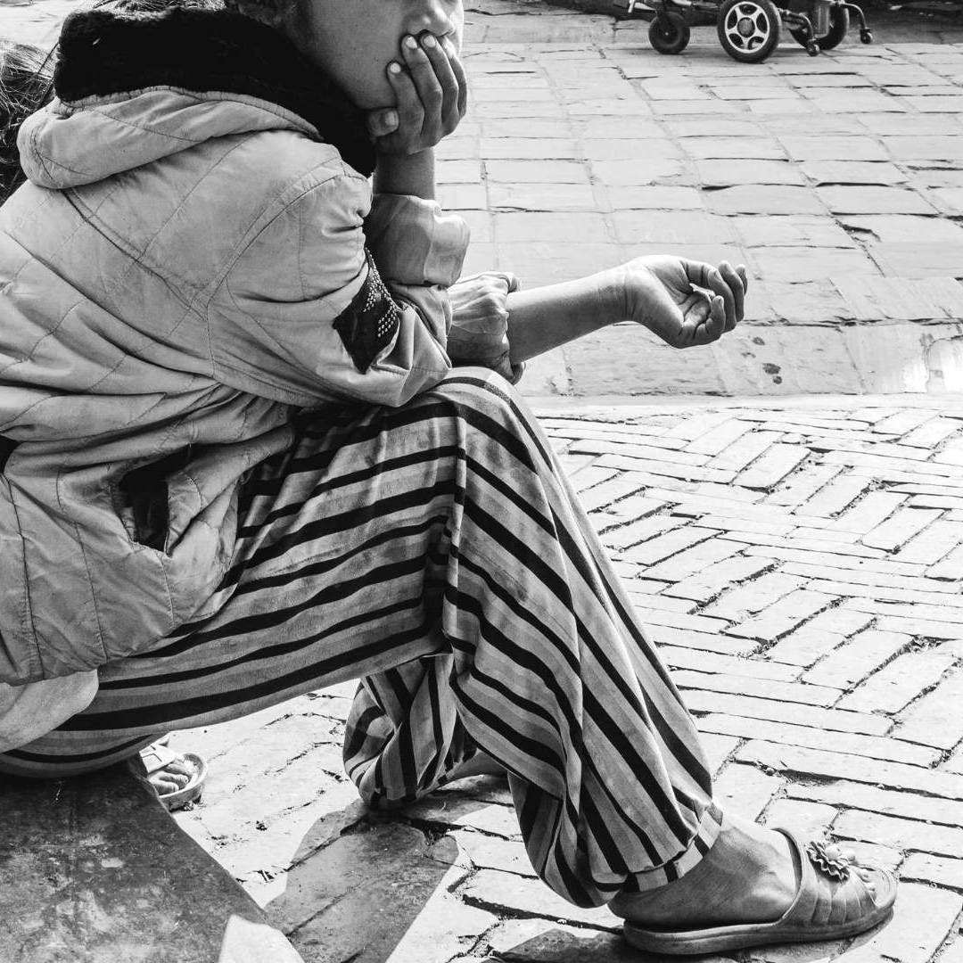 A black-and-white photo of two people sitting on a cobblestone street, facing away. One wears a striped outfit, appearing contemplative.