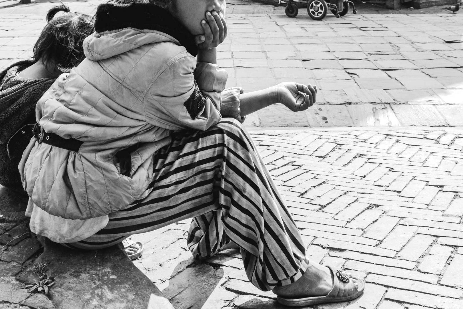 A black-and-white photo of two people sitting on a cobblestone street, facing away. One wears a striped outfit, appearing contemplative.