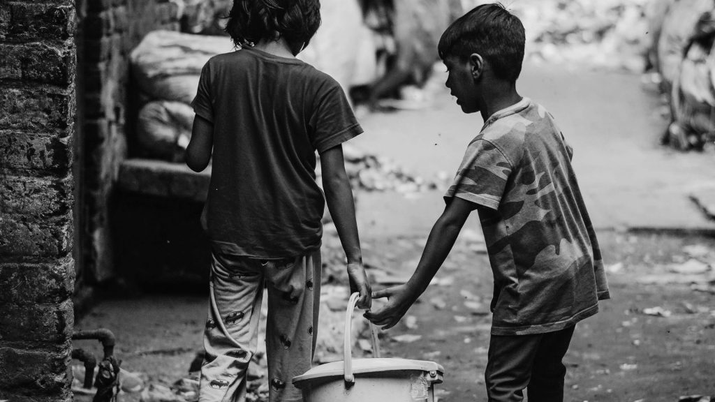 In a black and white photo, two young boys walk down an alley, one carrying a plastic bucket. The setting feels somber and conveys a mood of hardship.