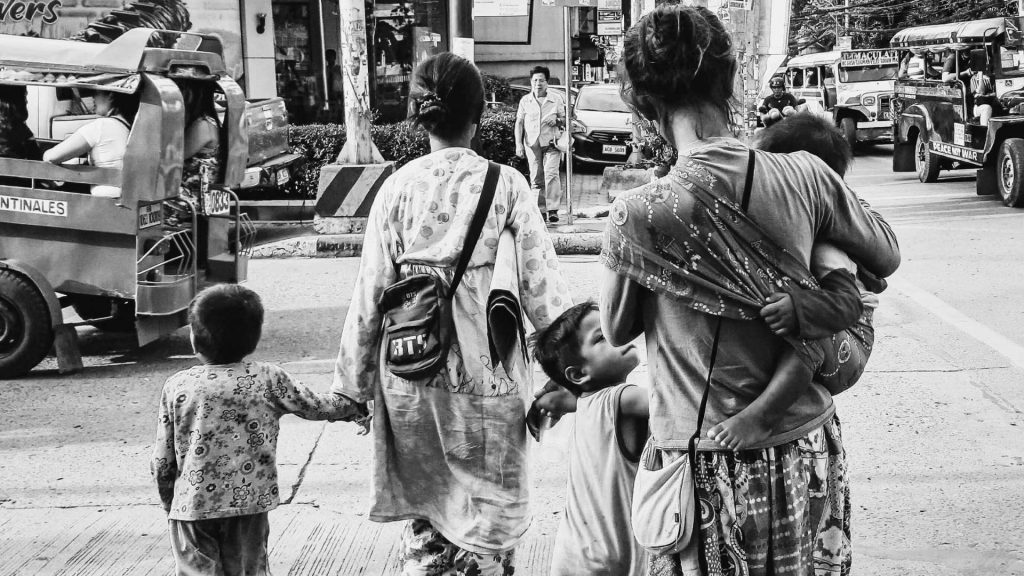 A black-and-white street scene shows two women with three children. One woman carries a baby, while a child holds her hand. The environment is urban, with vehicles and pedestrians. The mood appears busy and communal.