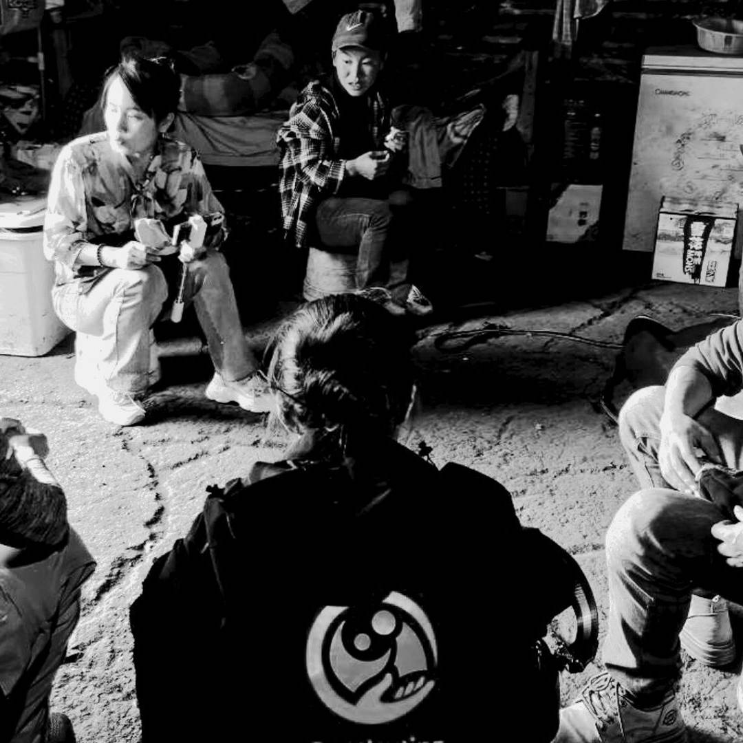 A group of five people sit in a circle in a dimly lit room. One person holds a baby. The atmosphere appears calm and focused, fostering conversation.