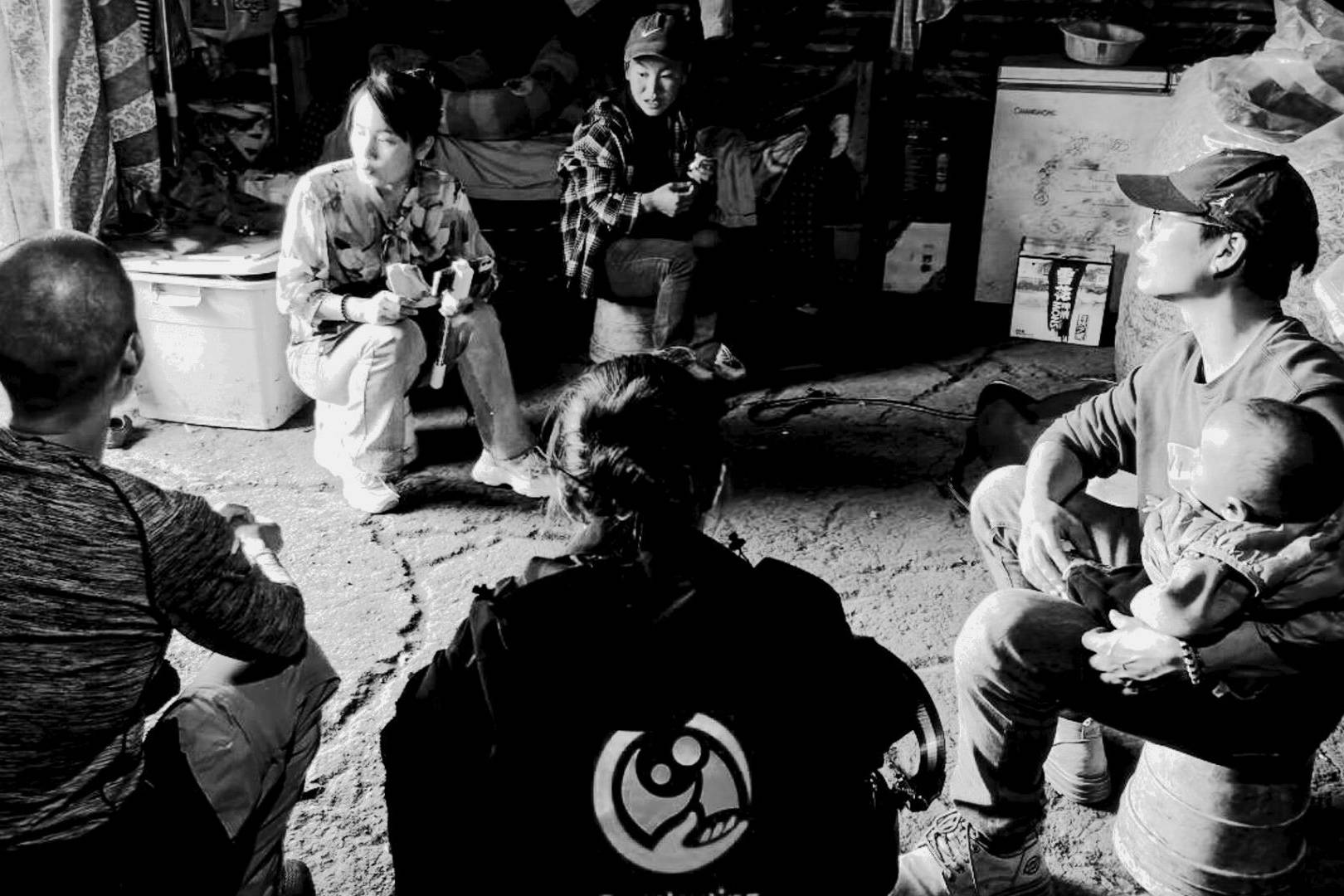A group of five people sit in a circle in a dimly lit room. One person holds a baby. The atmosphere appears calm and focused, fostering conversation.