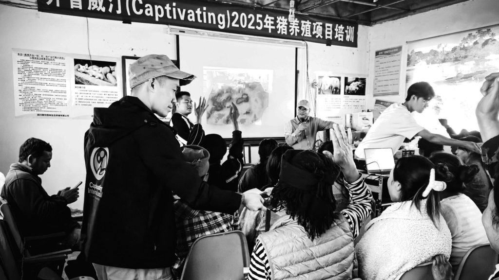 A group of people attentively participates in a workshop, with a speaker at the front by a projector screen and a person passing out materials. The mood is engaged and focused.