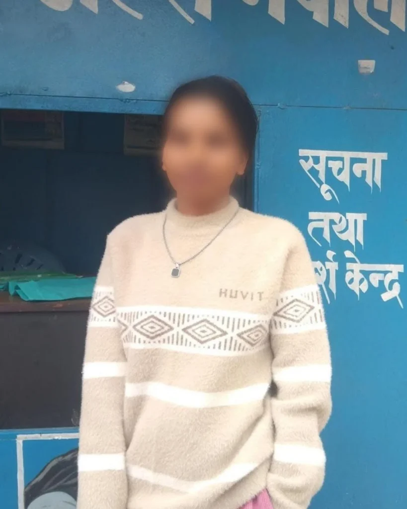 A person stands in front of a blue information booth, wearing a beige sweater with geometric patterns. White Hindi text is visible in the background. Her face blurred for privacy.