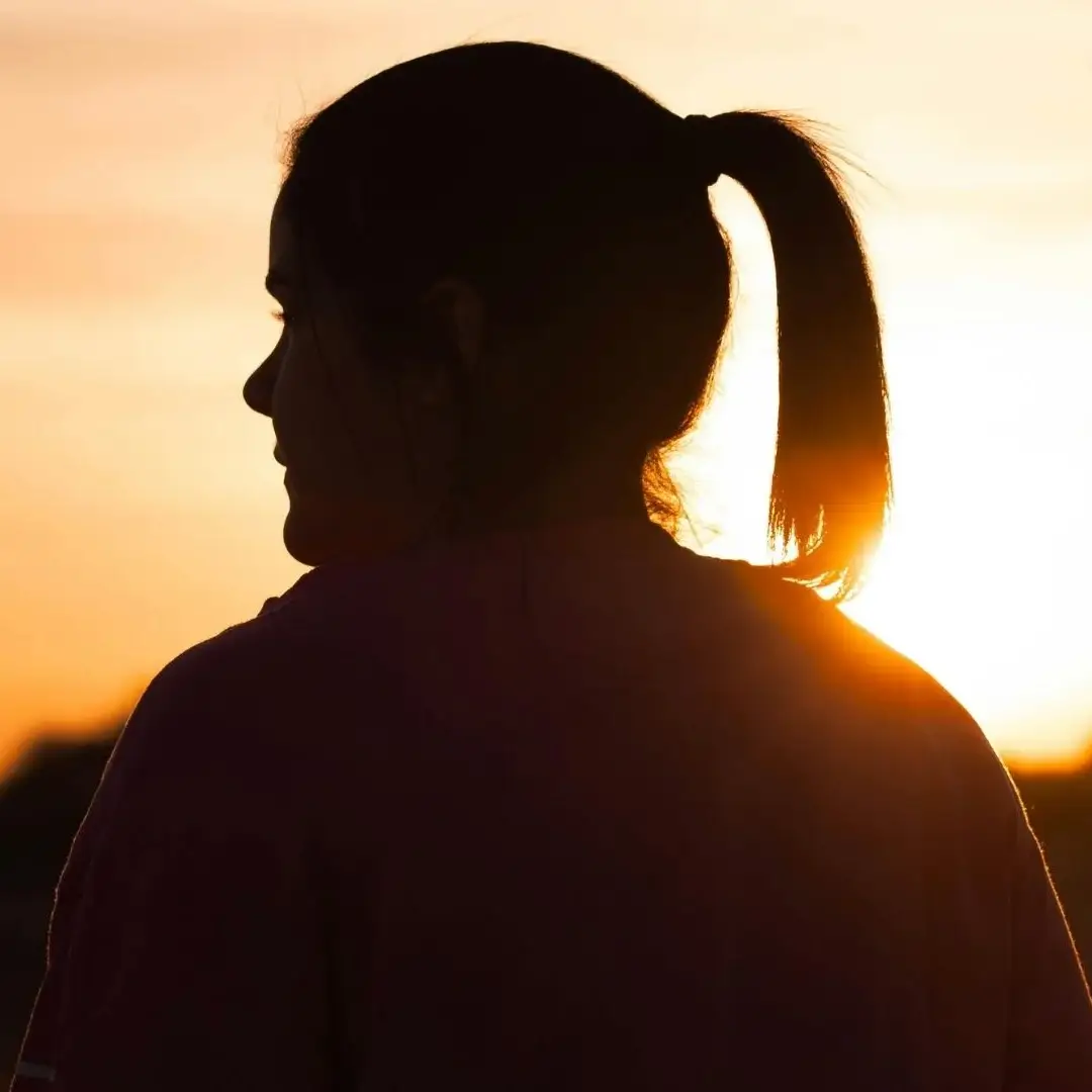 Silhouetted woman with a ponytail facing sunset, warm orange and yellow hues fill the sky, evoking a calm, contemplative atmosphere.