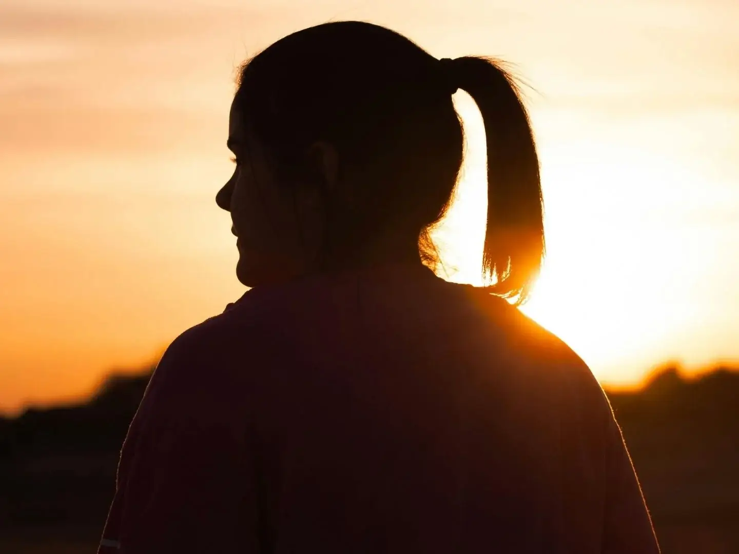 Silhouetted woman with a ponytail facing sunset, warm orange and yellow hues fill the sky, evoking a calm, contemplative atmosphere.