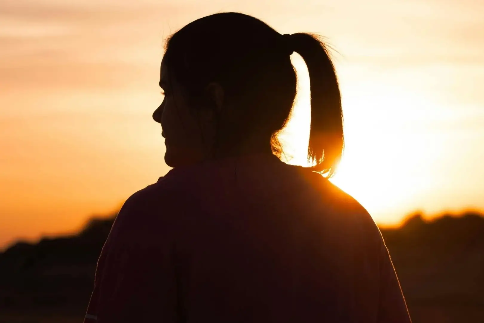 Silhouetted woman with a ponytail facing sunset, warm orange and yellow hues fill the sky, evoking a calm, contemplative atmosphere.
