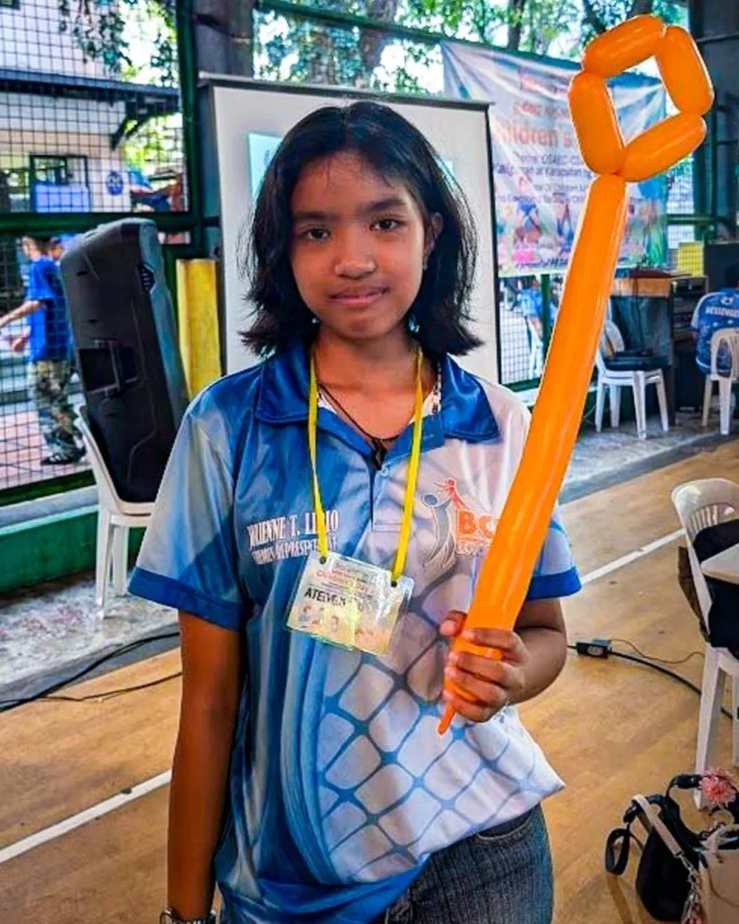 Young person in a blue sports shirt holds an orange balloon sculpture indoors. The setting is lively, possibly at an event or workshop.