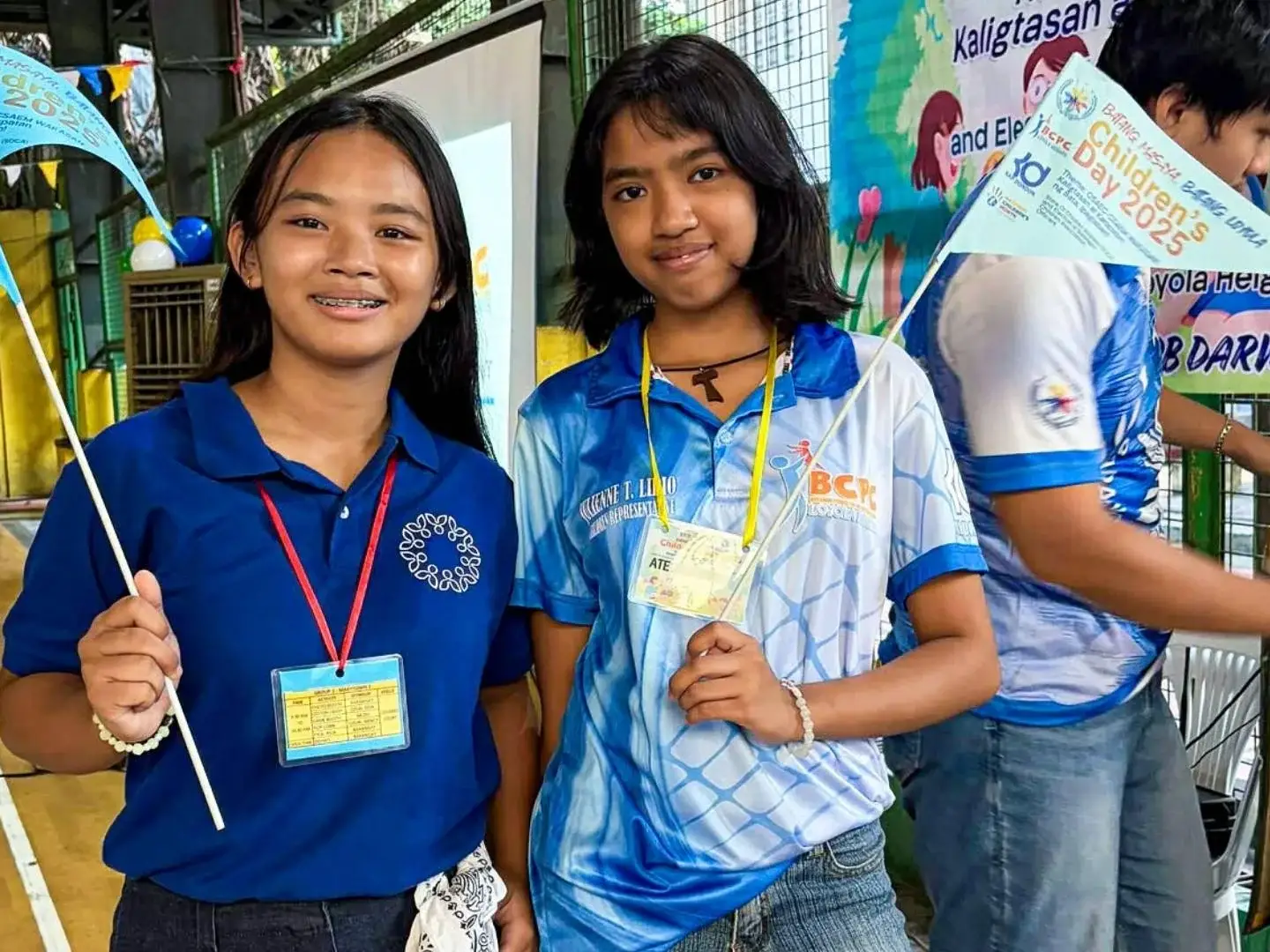 Two smiling girls holding blue flags pose together at an indoor event. They wear name tags and casual outfits, creating a joyful and lively atmosphere.