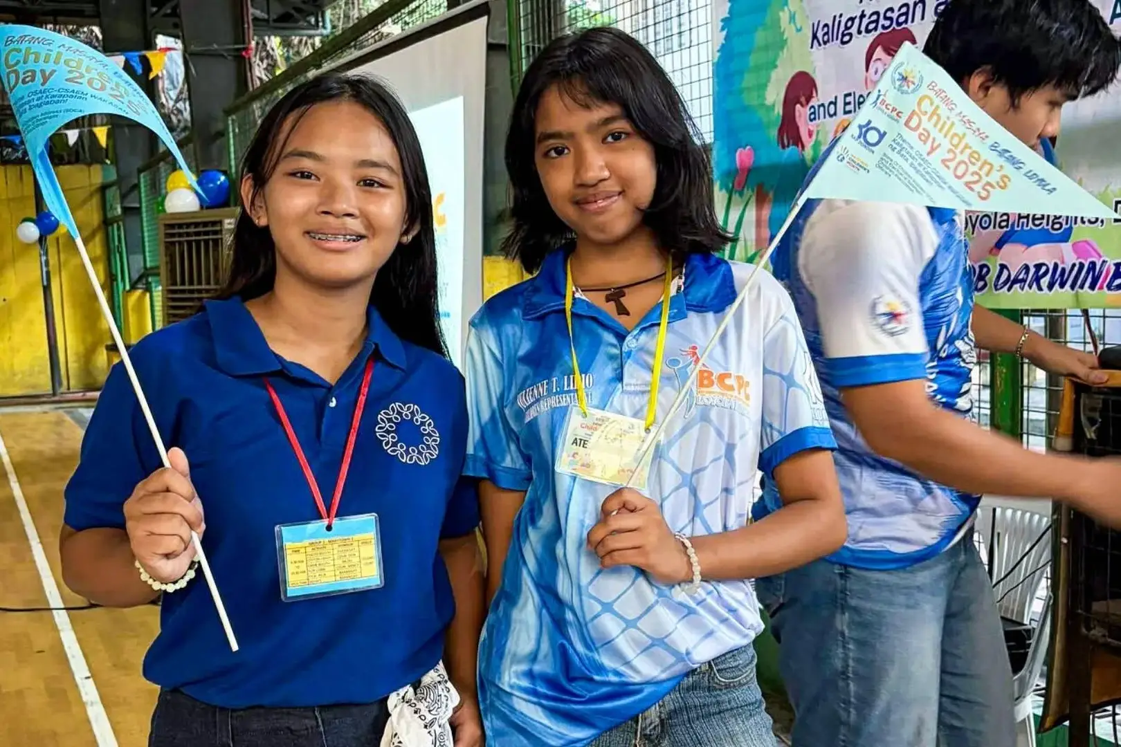 Two smiling girls holding blue flags pose together at an indoor event. They wear name tags and casual outfits, creating a joyful and lively atmosphere.