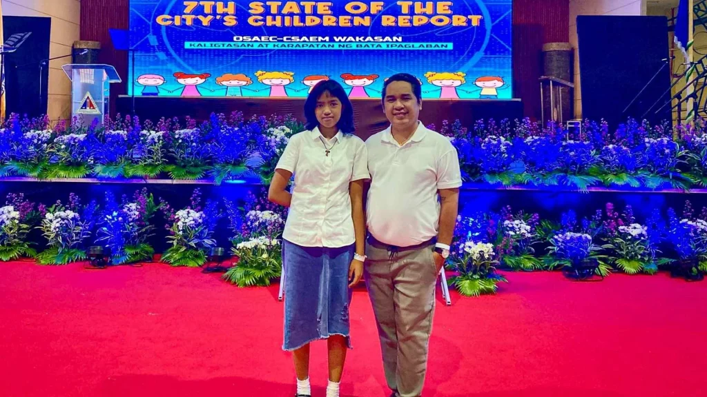 A smiling man and woman stand on a red carpet in front of a stage with vibrant purple flowers. Above them is a blue banner for the 7th State of the City's Children Report.