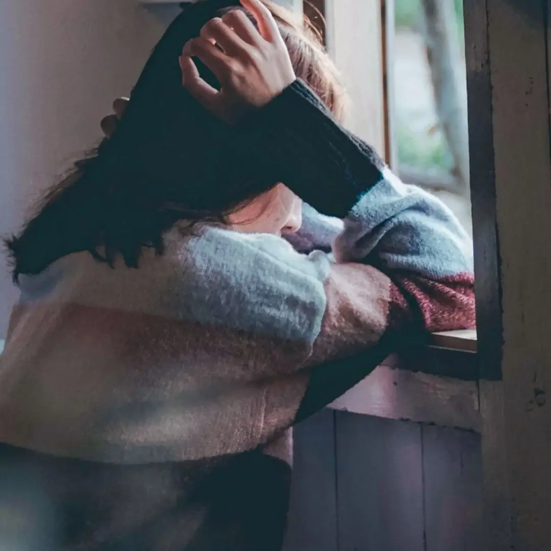 A person in a colorful sweater leans on a windowsill, gazing outside with a pensive expression. The scene conveys a mood of contemplation and solitude.
