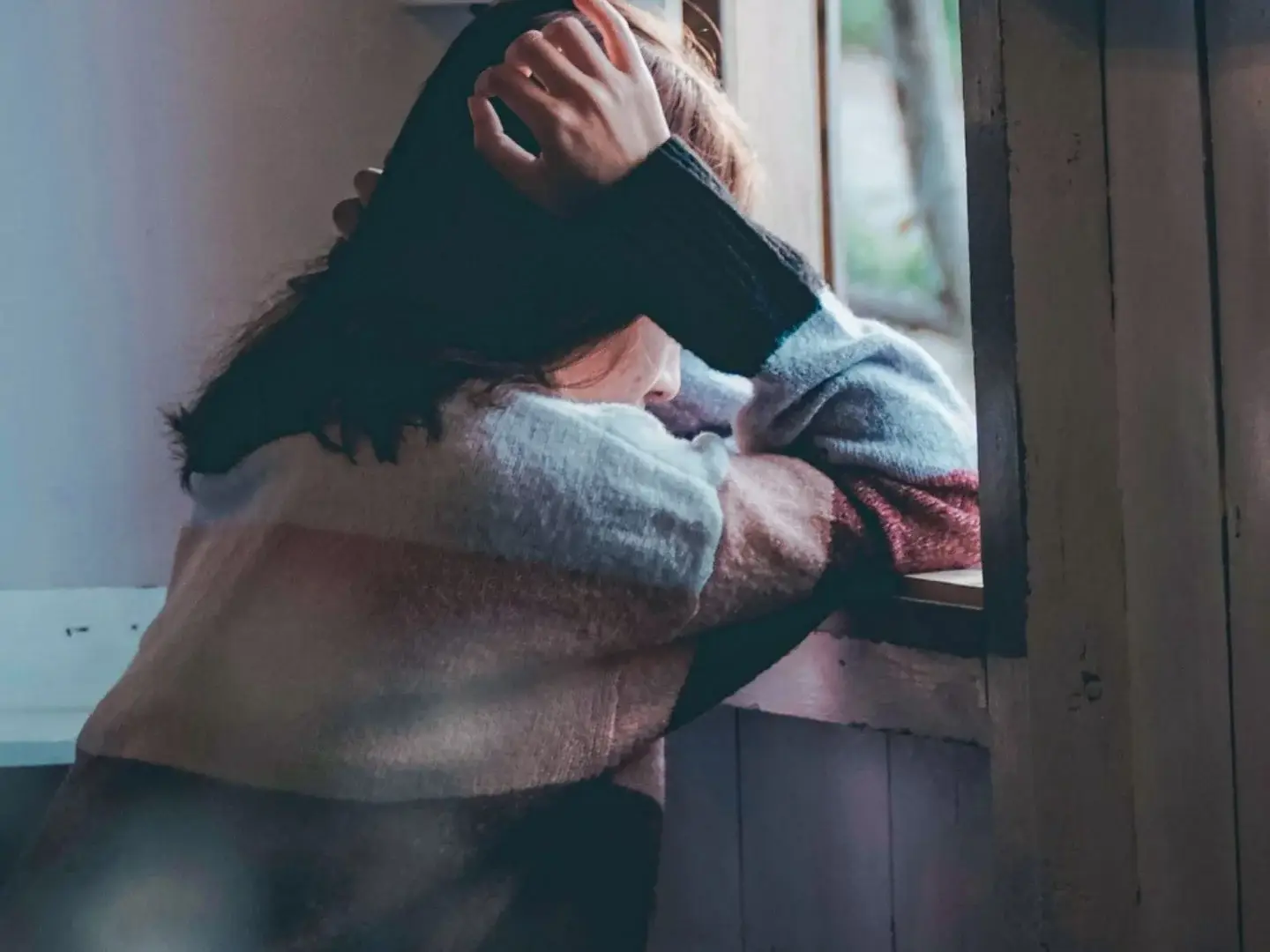 A person in a colorful sweater leans on a windowsill, gazing outside with a pensive expression. The scene conveys a mood of contemplation and solitude.