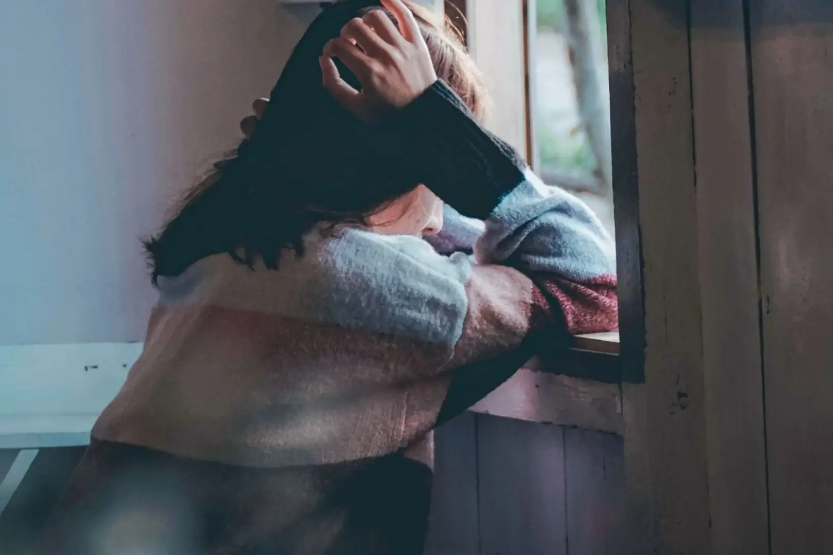 A person in a colorful sweater leans on a windowsill, gazing outside with a pensive expression. The scene conveys a mood of contemplation and solitude.