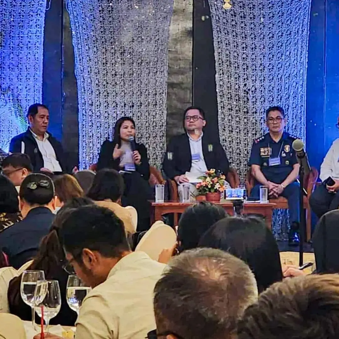 A panel of seven people sits on stage at a conference, engaged in discussion. The audience listens attentively. Blue lighting and floral decor set a formal tone.