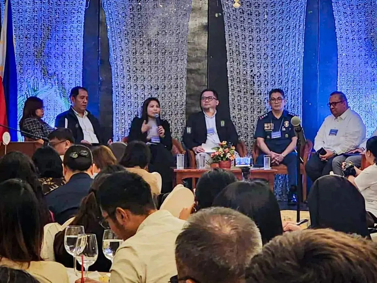 A panel of seven people sits on stage at a conference, engaged in discussion. The audience listens attentively. Blue lighting and floral decor set a formal tone.