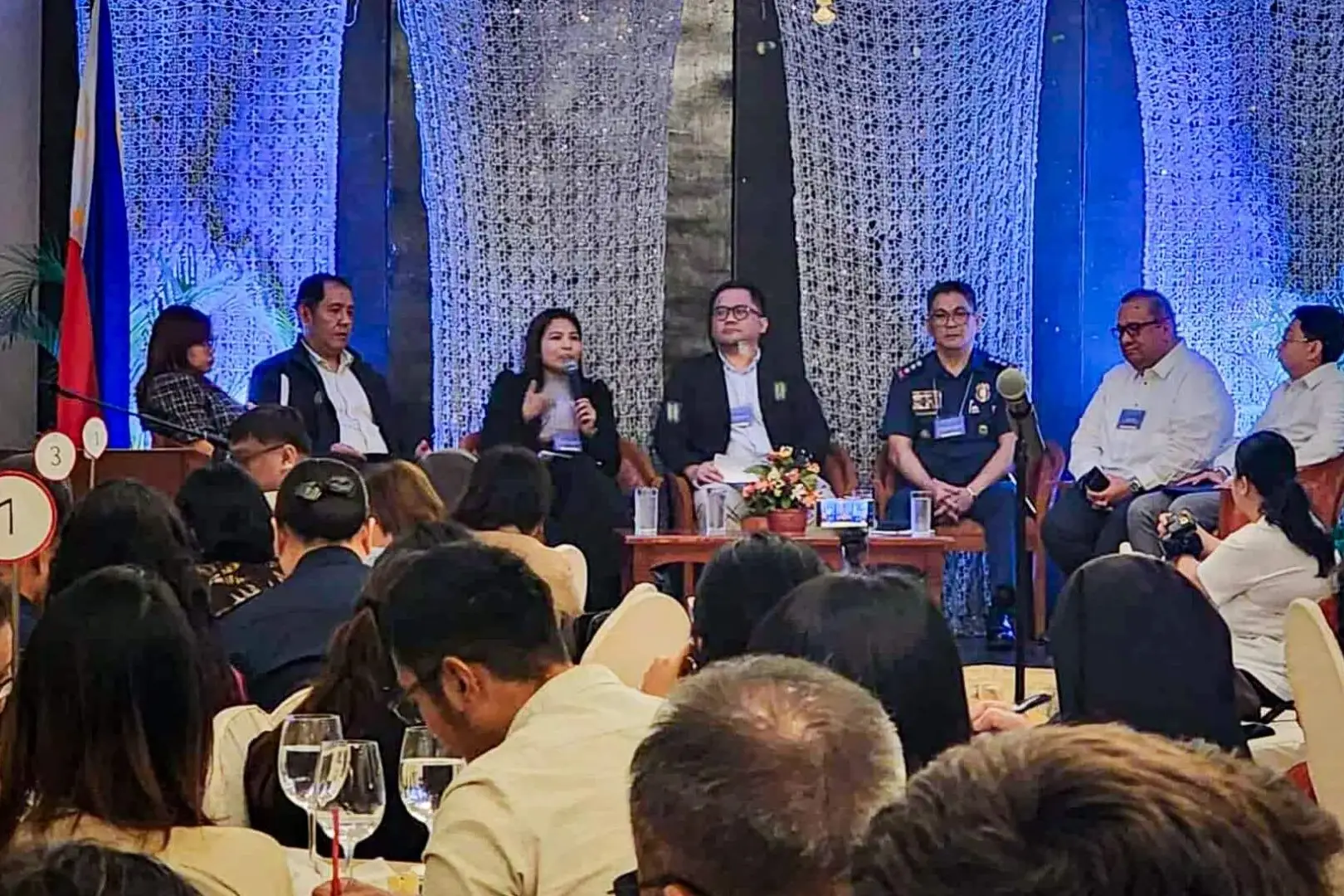 A panel of seven people sits on stage at a conference, engaged in discussion. The audience listens attentively. Blue lighting and floral decor set a formal tone.