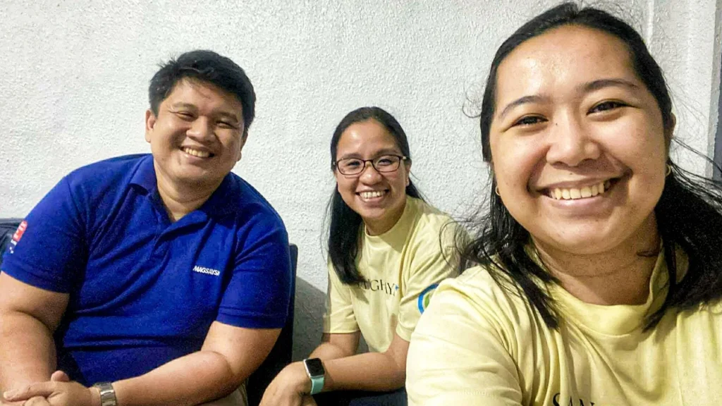 Three people smiling, two wearing yellow shirts and one in blue, pose for a selfie against a plain white wall, creating a cheerful atmosphere.
