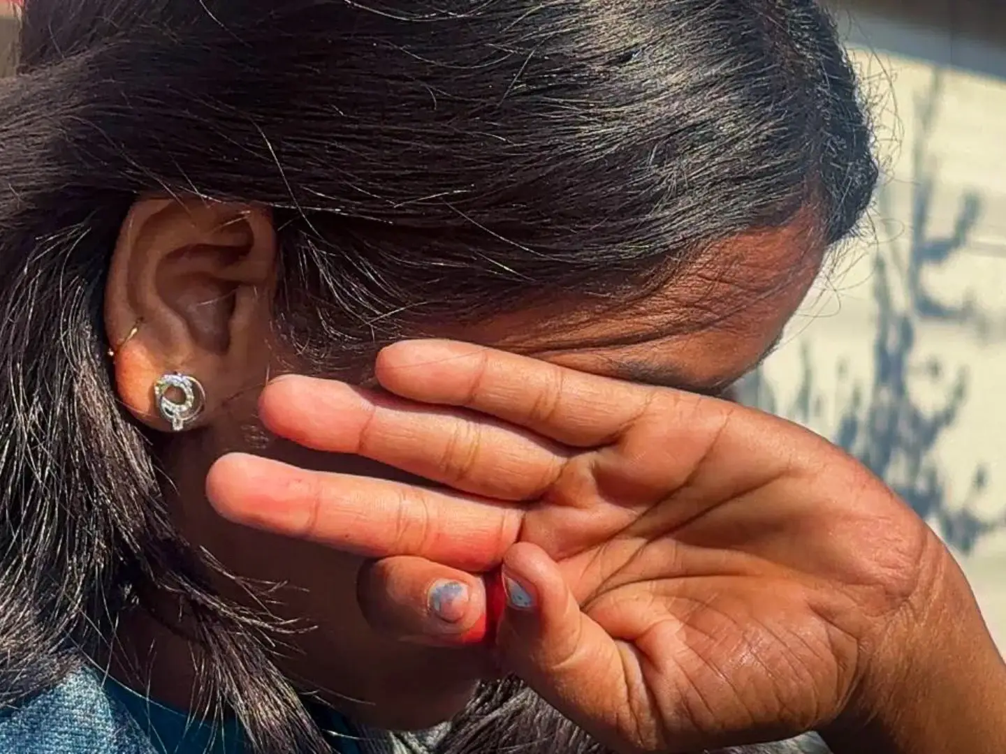 A person with long, dark hair covers their face with their hands, conveying shyness or distress. Sunlight casts shadows on their hair and a background wall.