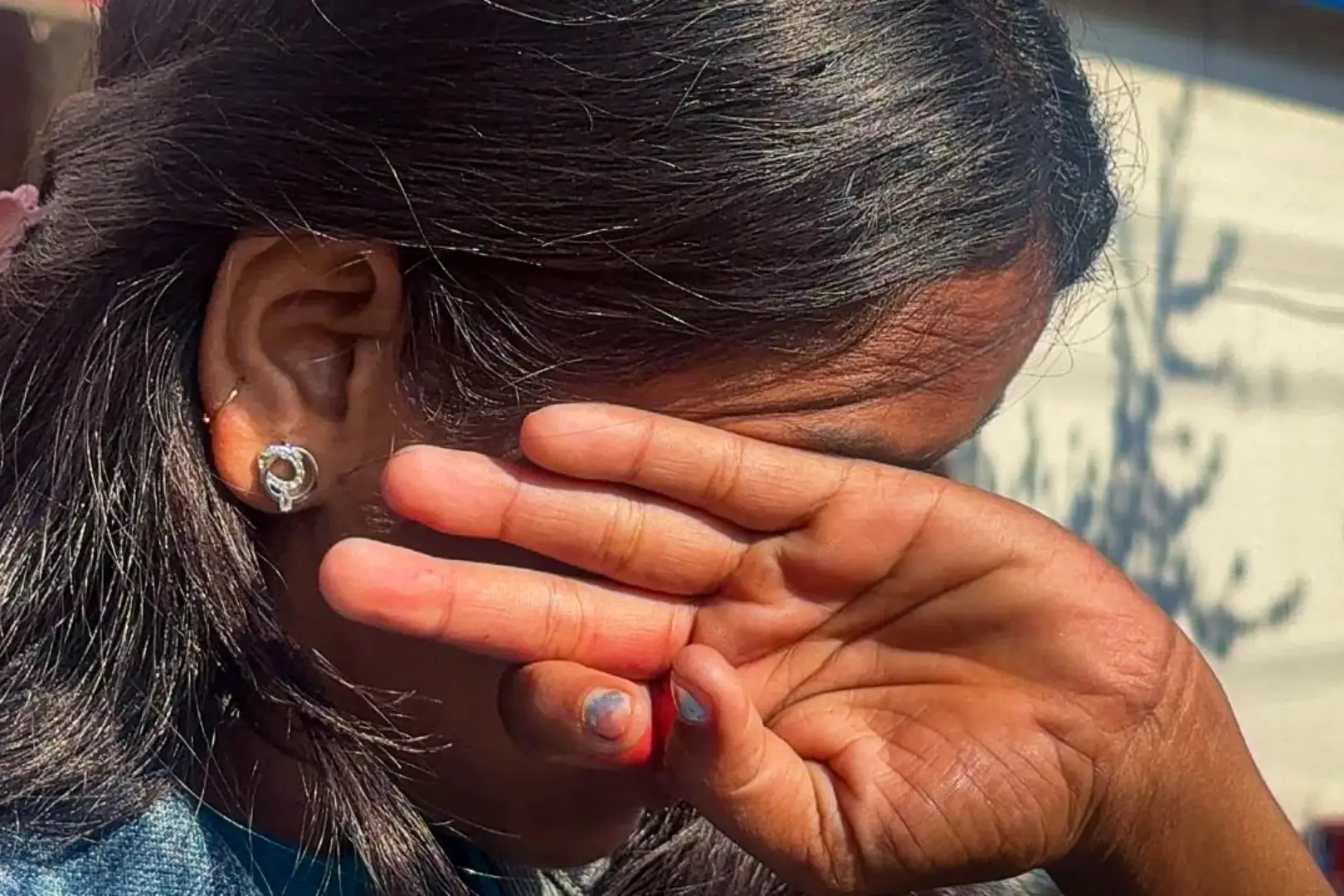 A person with long, dark hair covers their face with their hands, conveying shyness or distress. Sunlight casts shadows on their hair and a background wall.