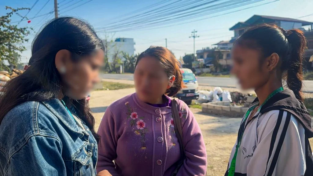 Three women stand in conversation outdoors under a clear blue sky, near a street with houses in the background. Their faces blurred for privacy.