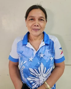 A middle-aged Filipino woman wearing a blue and white collared shirt smiles softly. She stands against a plain beige background, conveying a warm and friendly demeanor.
