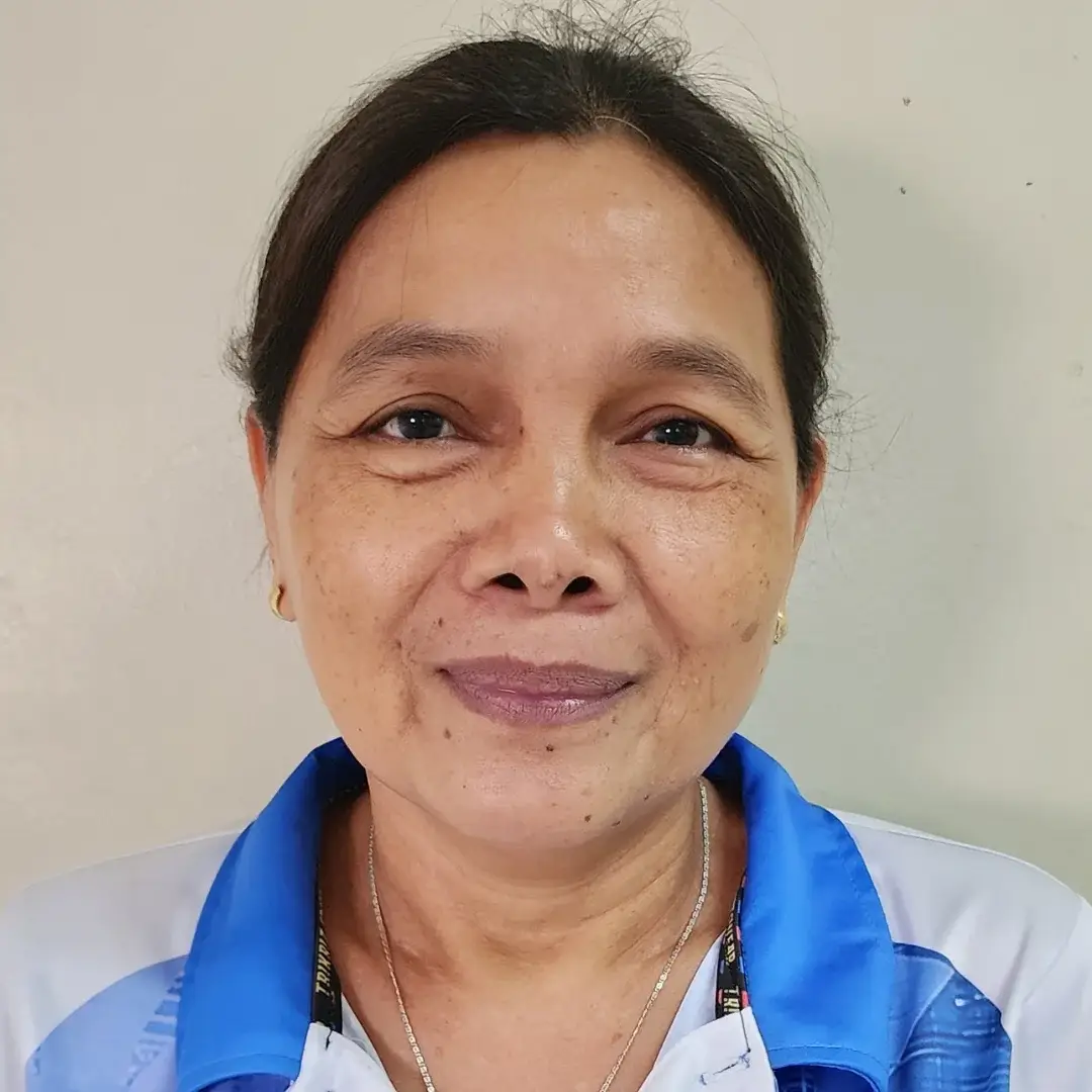 A middle-aged Filipino woman wearing a blue and white collared shirt smiles softly. She stands against a plain beige background, conveying a warm and friendly demeanor.