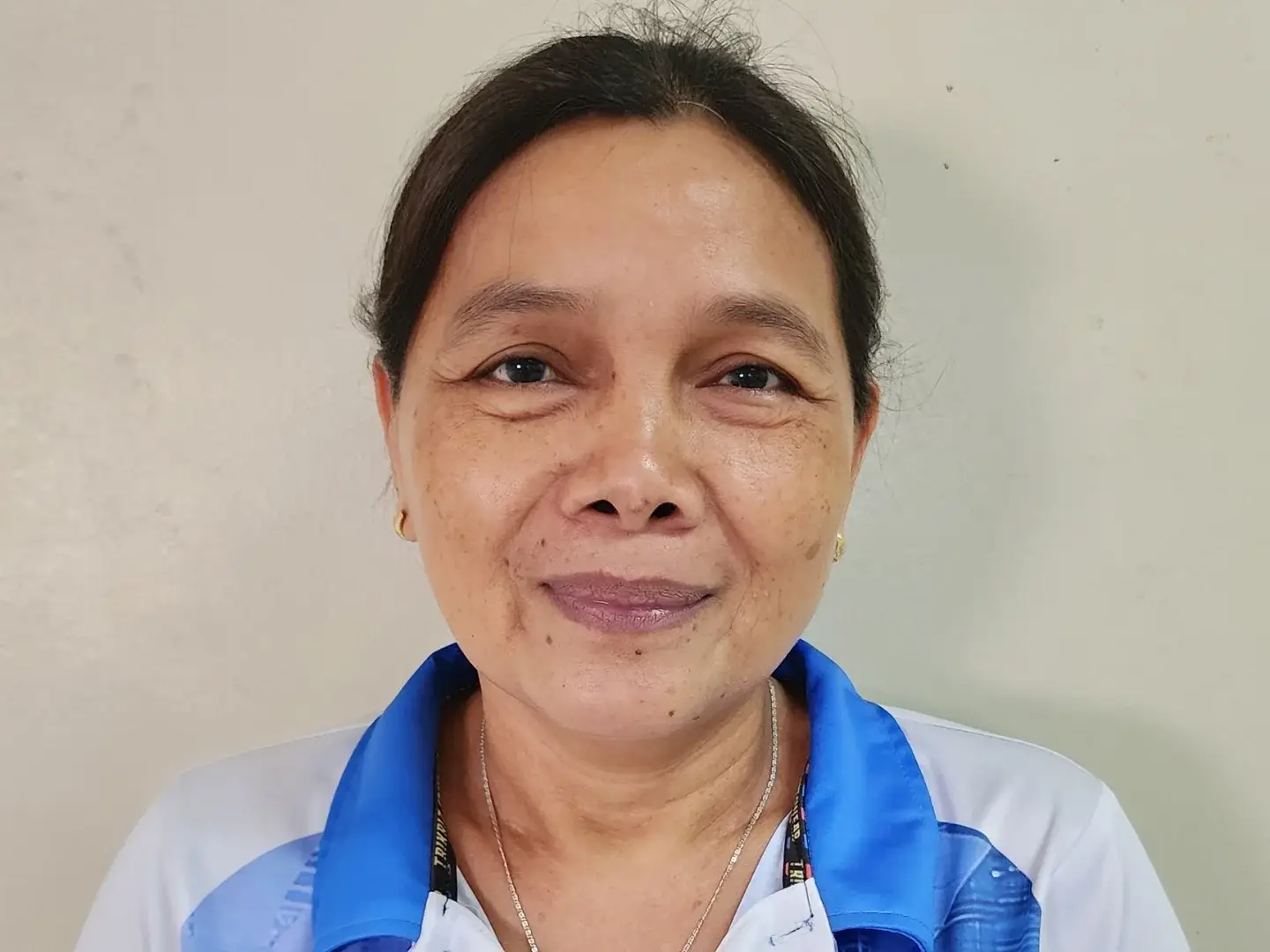 A middle-aged Filipino woman wearing a blue and white collared shirt smiles softly. She stands against a plain beige background, conveying a warm and friendly demeanor.