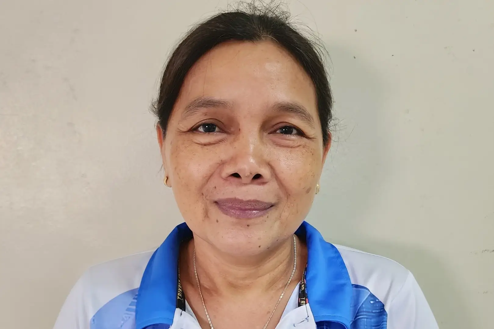 A middle-aged Filipino woman wearing a blue and white collared shirt smiles softly. She stands against a plain beige background, conveying a warm and friendly demeanor.