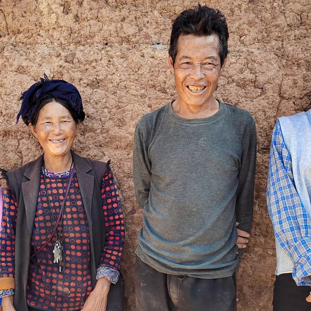 Four people stand smiling against a textured, earthen wall. They are dressed in casual, colorful clothing. The mood is warm and joyful.