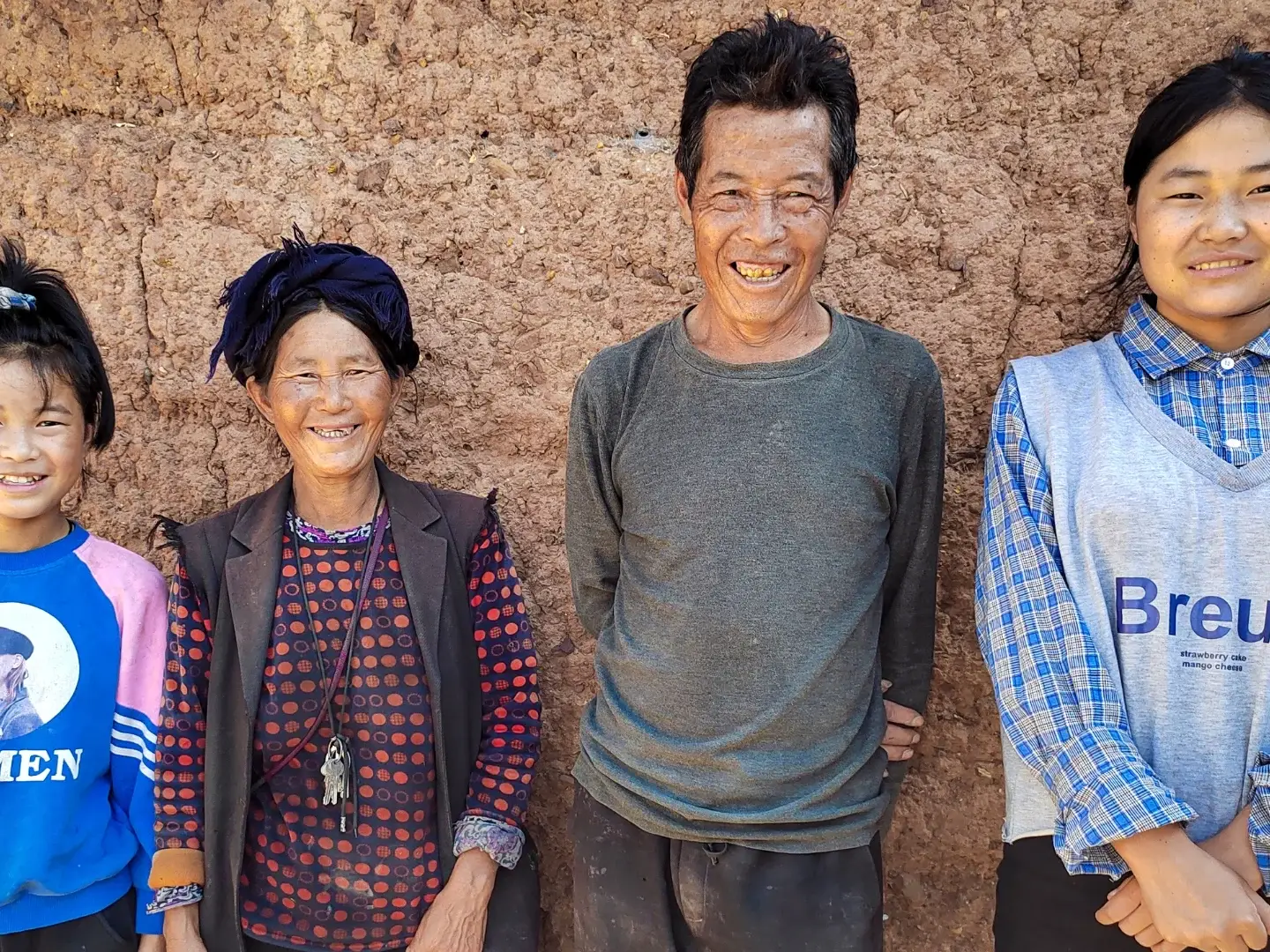 Four people stand smiling against a textured, earthen wall. They are dressed in casual, colorful clothing. The mood is warm and joyful.