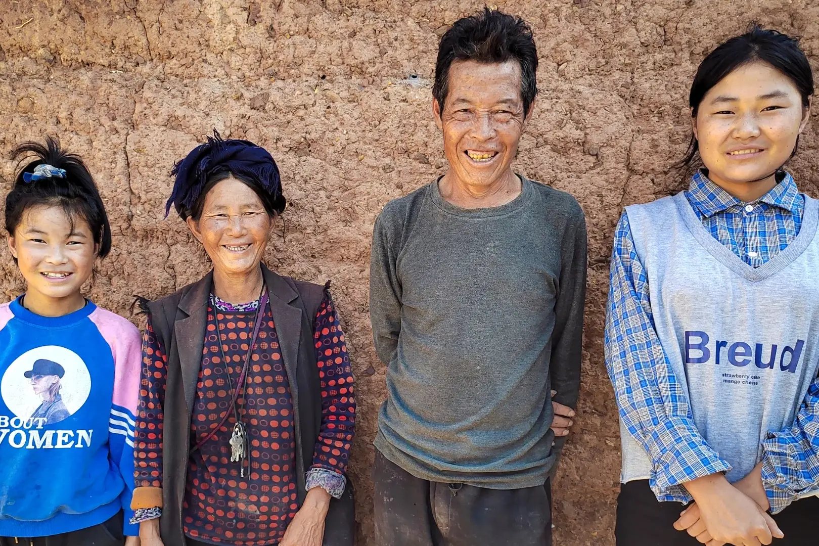 Four people stand smiling against a textured, earthen wall. They are dressed in casual, colorful clothing. The mood is warm and joyful.