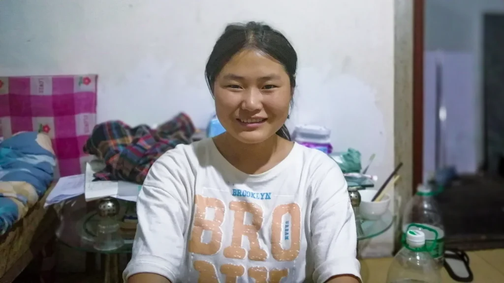 A woman smiles warmly in a cozy, cluttered room, wearing a white t-shirt. The background has colorful fabrics and bottles, conveying a homely feel.