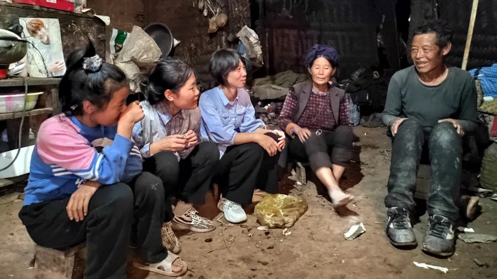 A group of five people sits in a dimly lit, rustic room, engaged in conversation. The mood appears warm and communal, highlighting togetherness.
