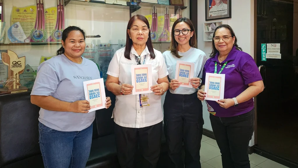 Four women stand in a Barangay office holding booklets, smiling. Trophy-filled cabinet and framed picture in the background convey achievement.